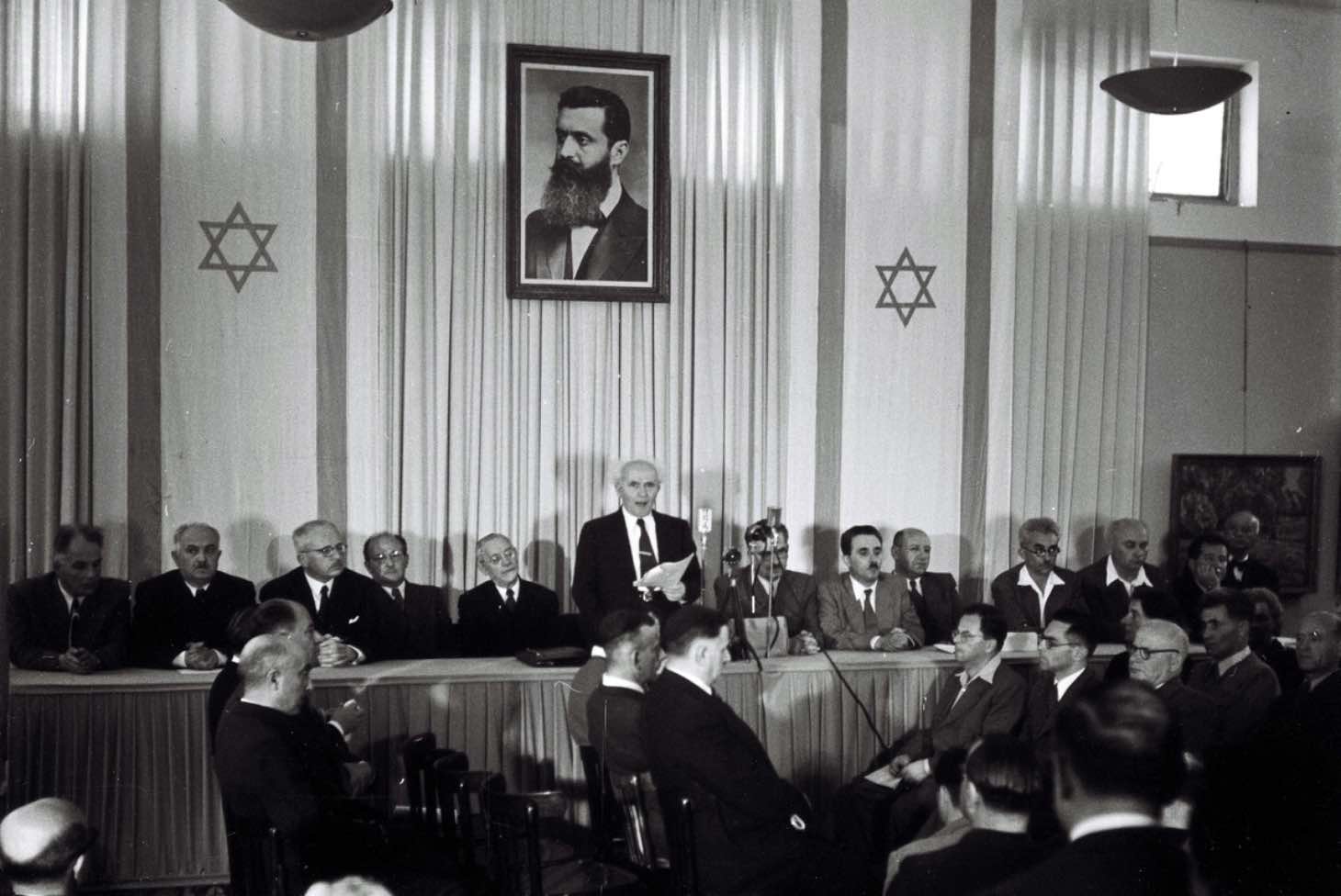 Israels første statsminister, David Ben Gurion, erklærer staten Israel for opprettet i 1948. Bildet på veggen er av Theodor Herzl, grunnlegger av den sionistiske bevegelse. SNL/Public Domain.